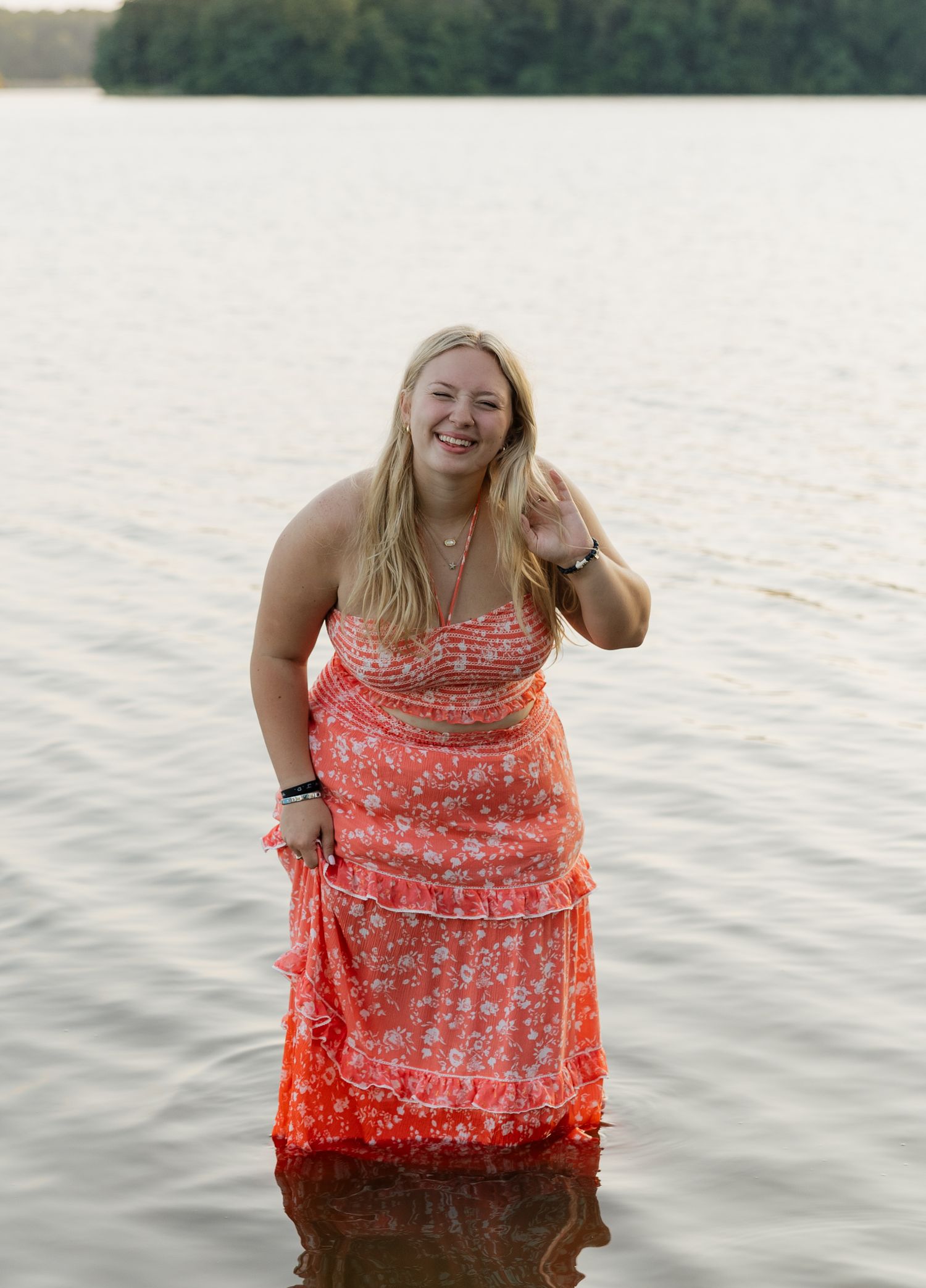 Someone wearing a coral floral dress stands in shallow water at sunset near a tree-lined shore.