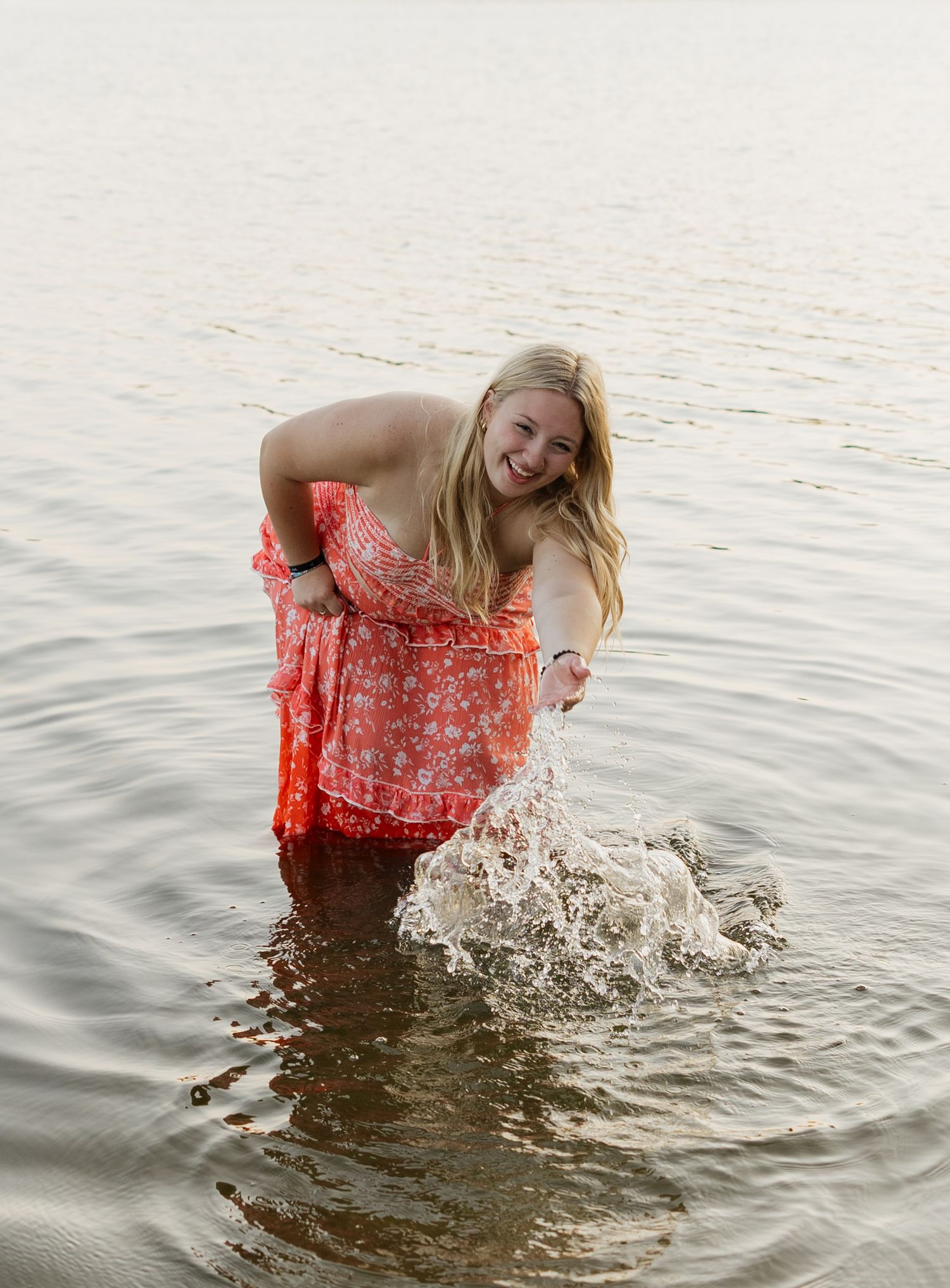 A playful splash sequence in a coral dress at the lake's edge during sunset.