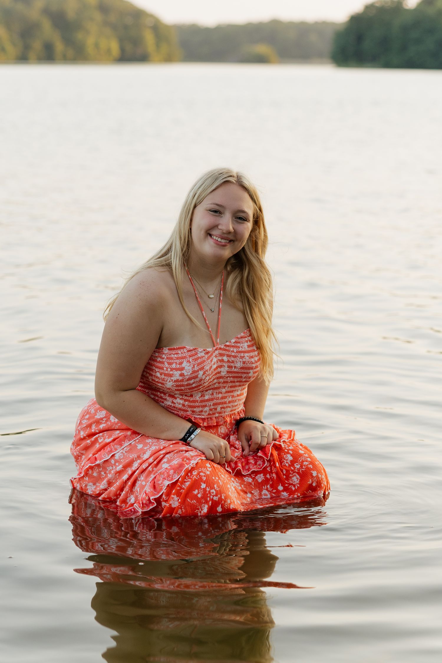 A person in a coral floral dress sits in calm lake water at sunset with trees visible in the background.