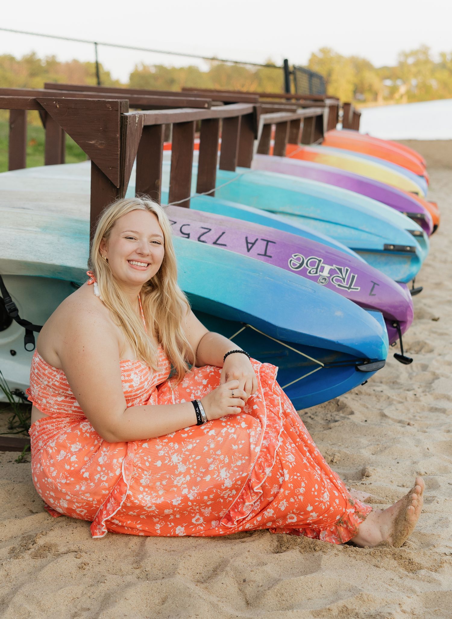 A colorful row of kayaks lines the beach while someone in a coral dress sits on the sand.