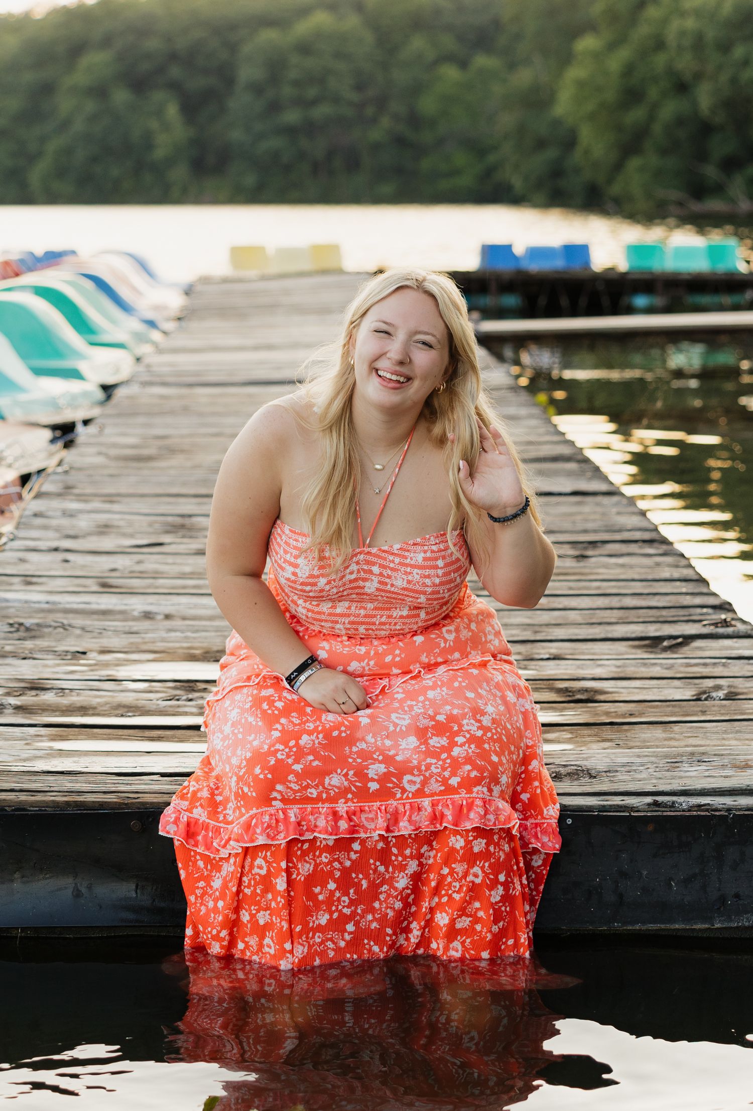 A joyful person in a coral floral dress sits on a wooden dock by the water with boats in the background.