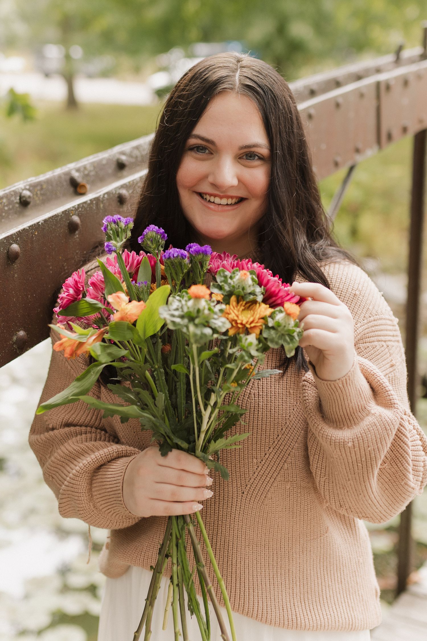 Someone in a cozy beige sweater holds a vibrant bouquet of colorful flowers while standing outdoors on a sunny day.