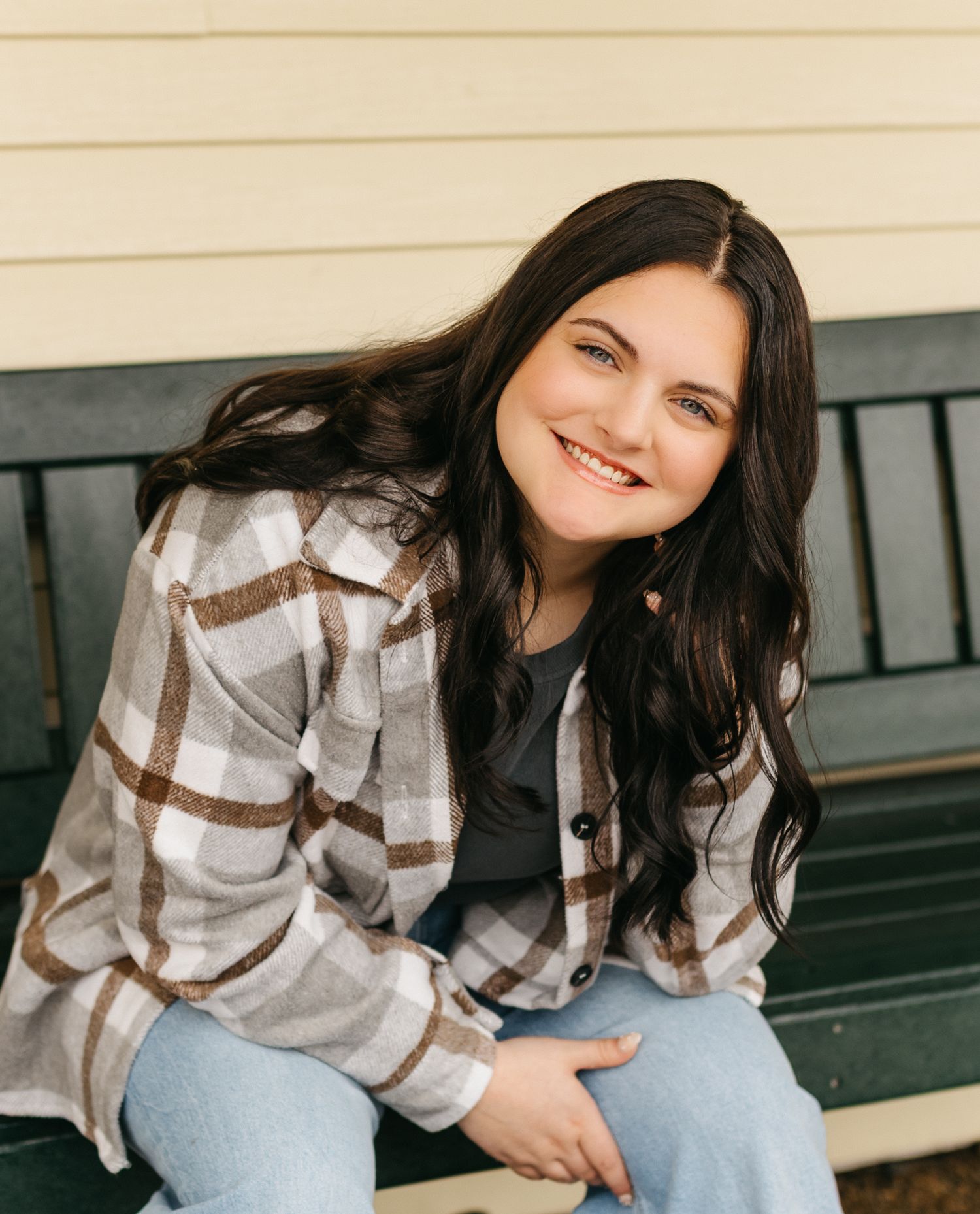 A person in a brown plaid flannel shirt and jeans sits smiling on a wooden bench outdoors.
