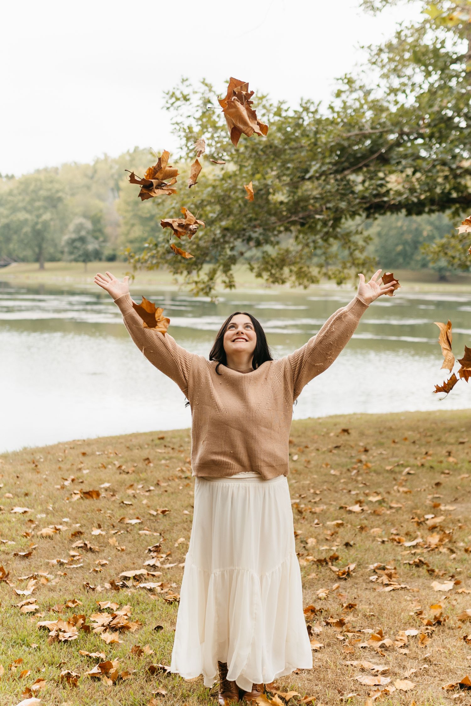 A person in a beige sweater and white dress joyfully throws autumn leaves by a lake.