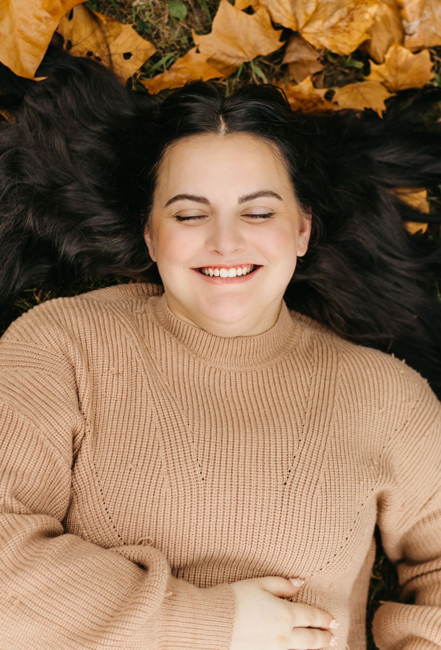 Person lying on autumn leaves wearing beige sweater with dark hair spread out in artistic portrait.