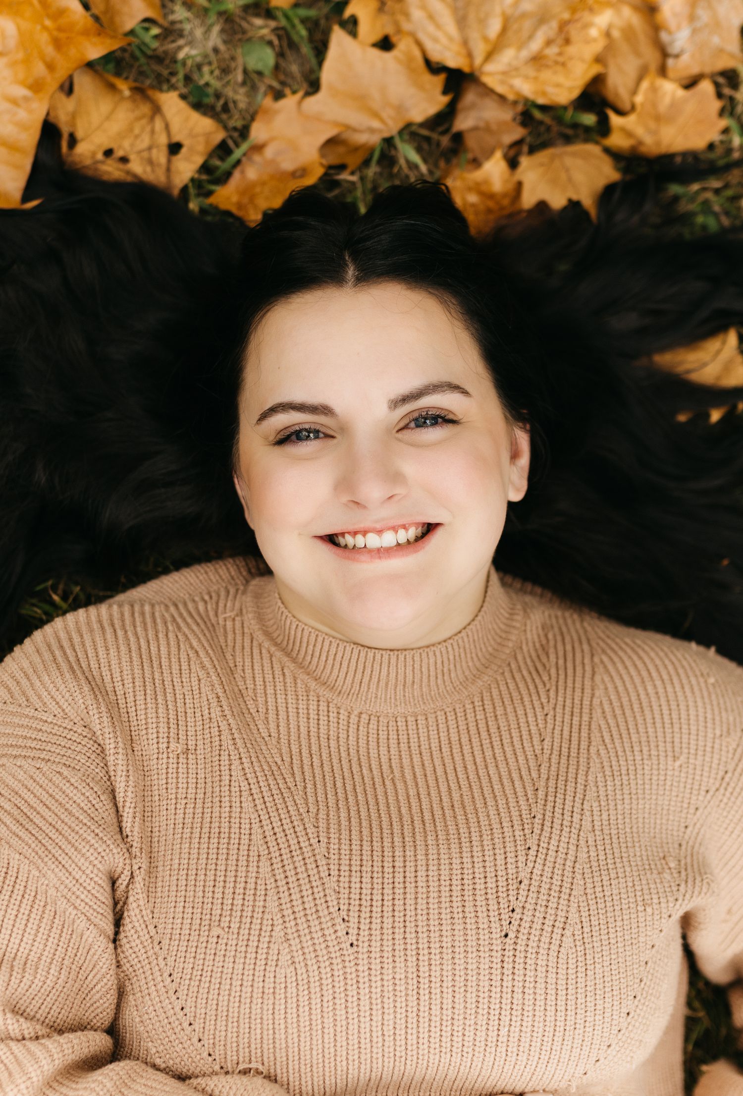Someone wearing a beige sweater smiles while lying on autumn leaves.