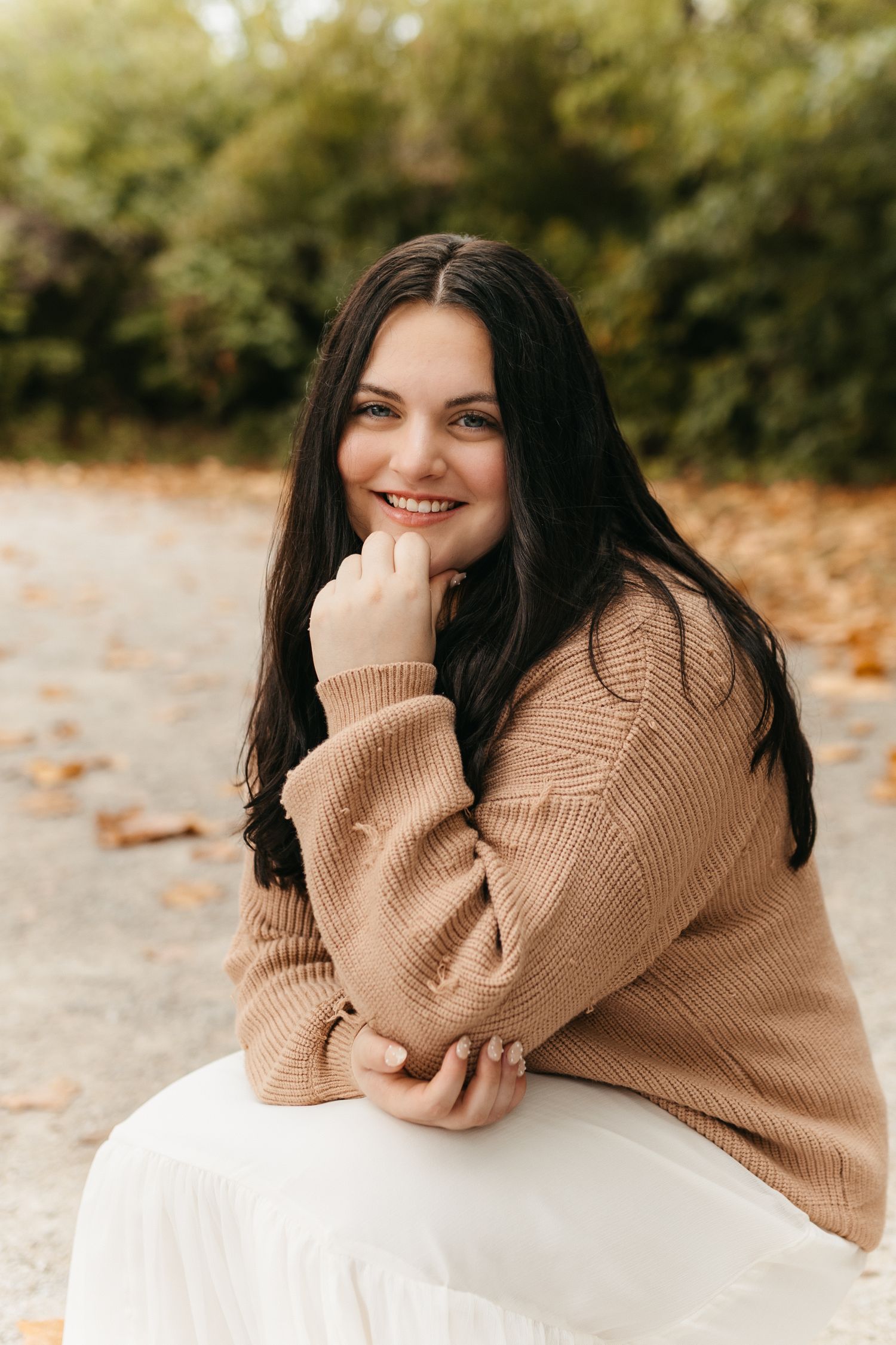 A person in a cozy beige sweater poses on a white surface surrounded by fall foliage.