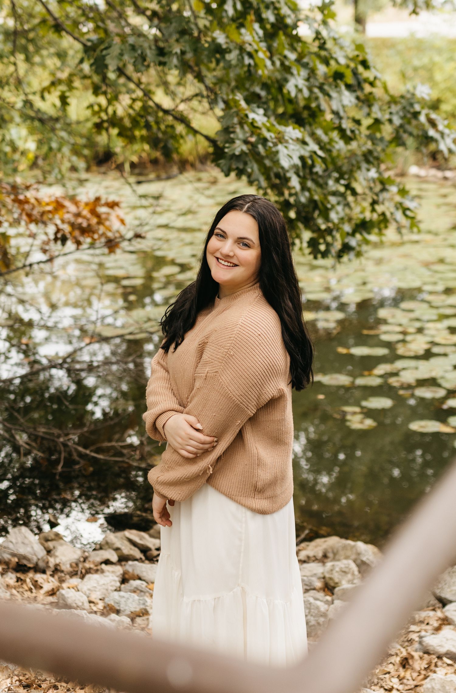 Person in beige sweater and white dress poses near pond with autumn foliage in background.