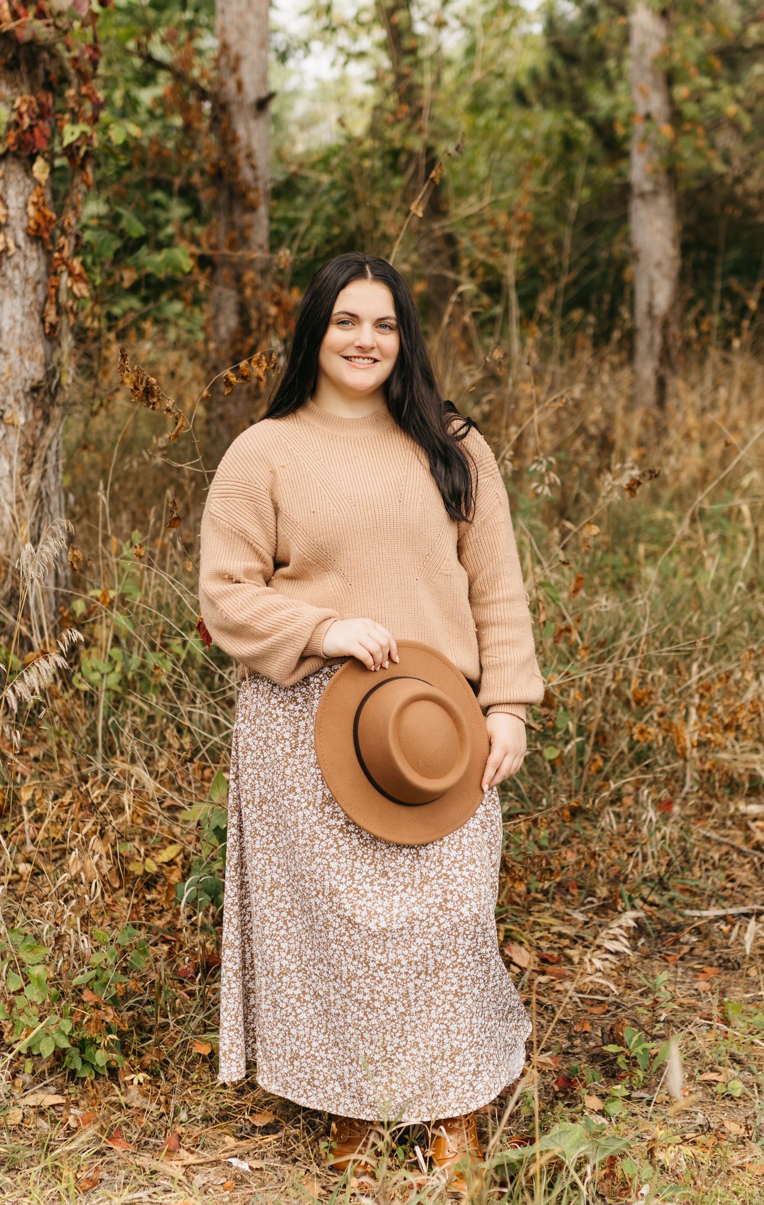 Autumn outdoor photoshoot featuring a floral dress and beige sweater with a brown fedora hat against fall foliage.