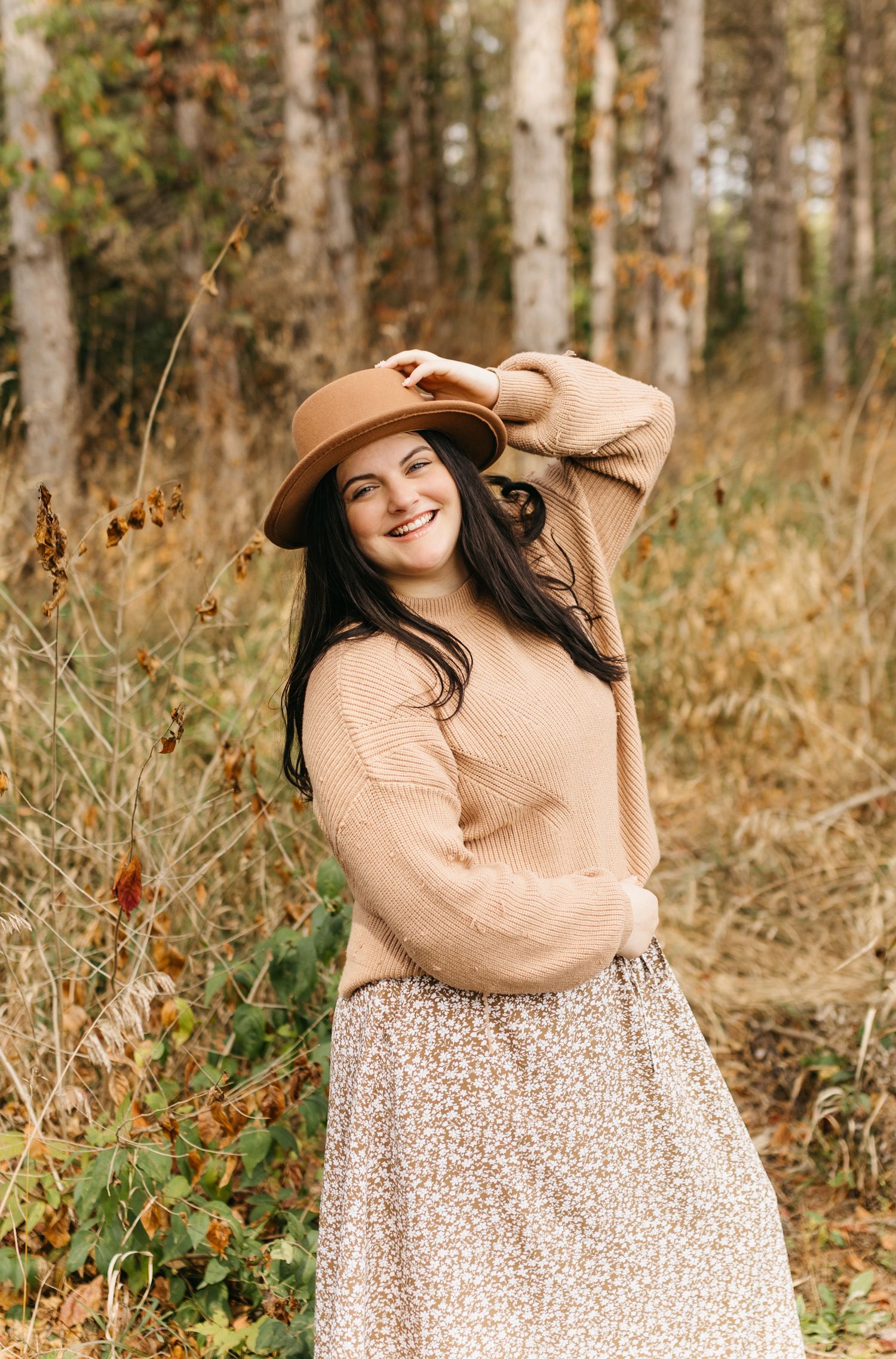 A person in a tan sweater and floral dress poses playfully in an autumn forest setting.