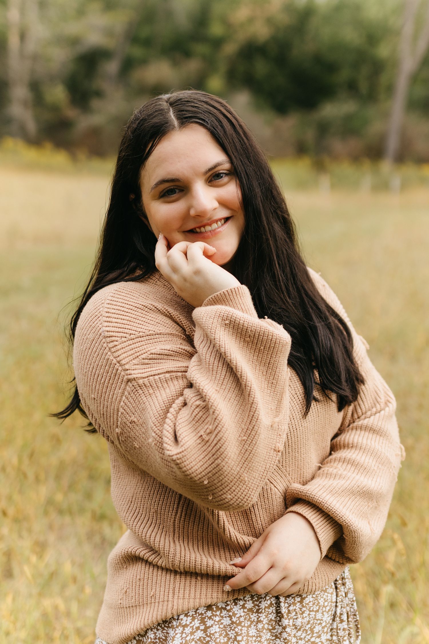 A person wearing a beige knit sweater poses playfully in an outdoor field setting with autumn grass.