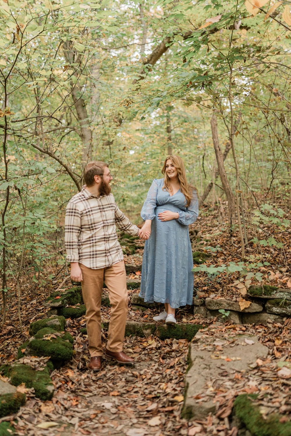 A couple walks together on a leaf-covered forest path during autumn, wearing plaid and blue outfits.