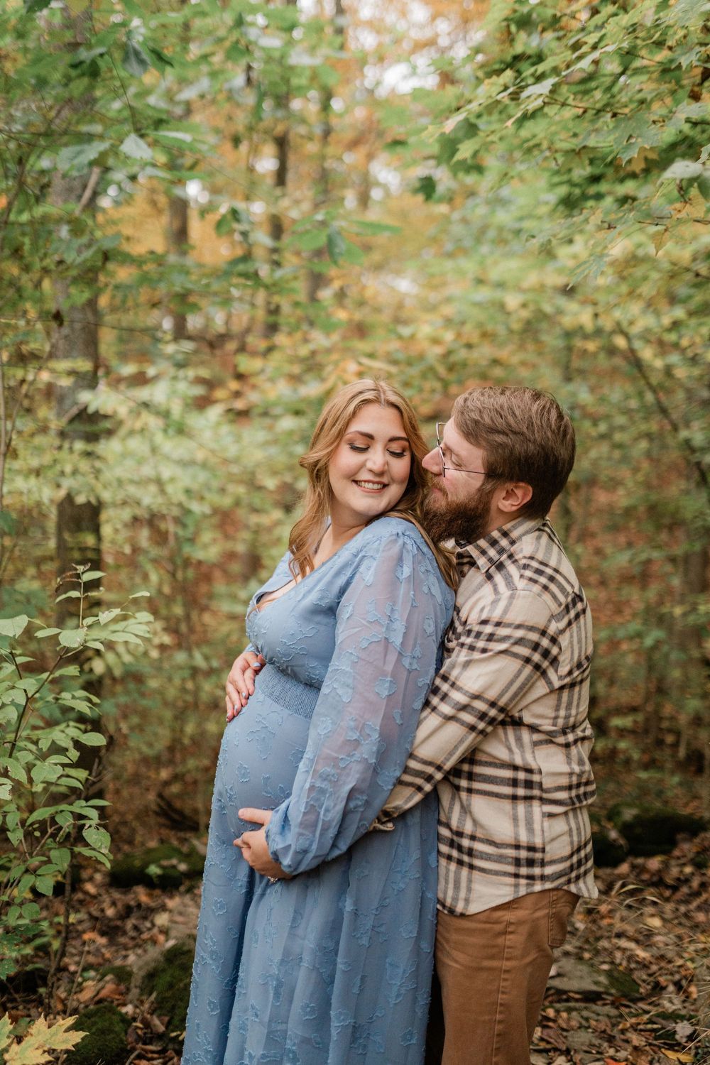 A romantic autumn maternity photo session shows a couple embracing in a wooded setting with fall foliage.