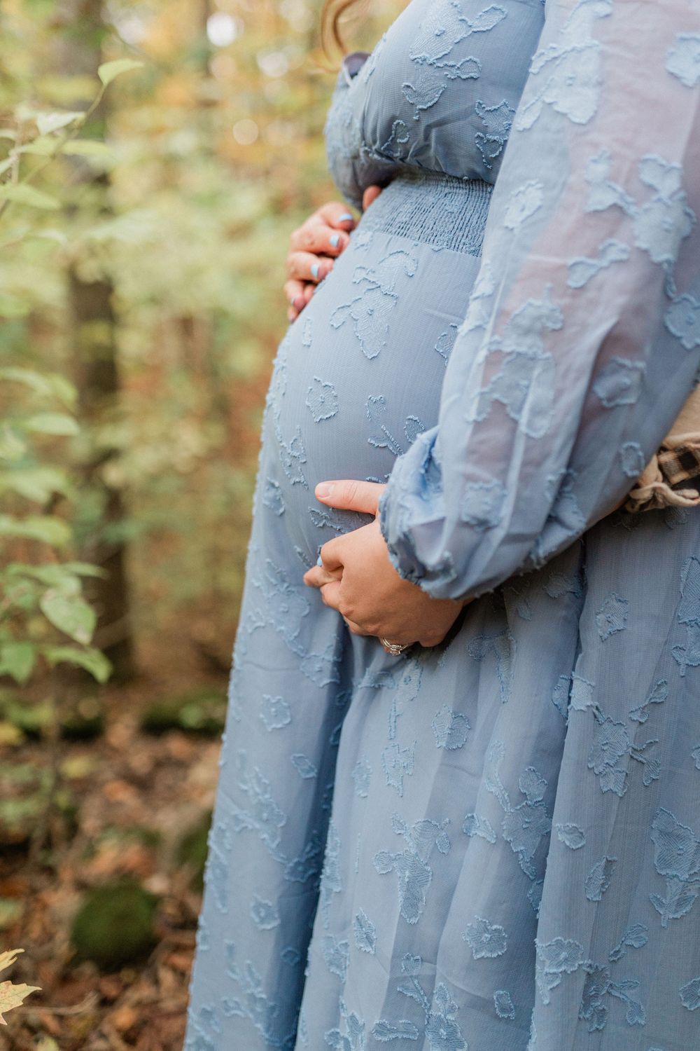Close-up details of a light blue lace maternity dress against autumn forest backdrop.