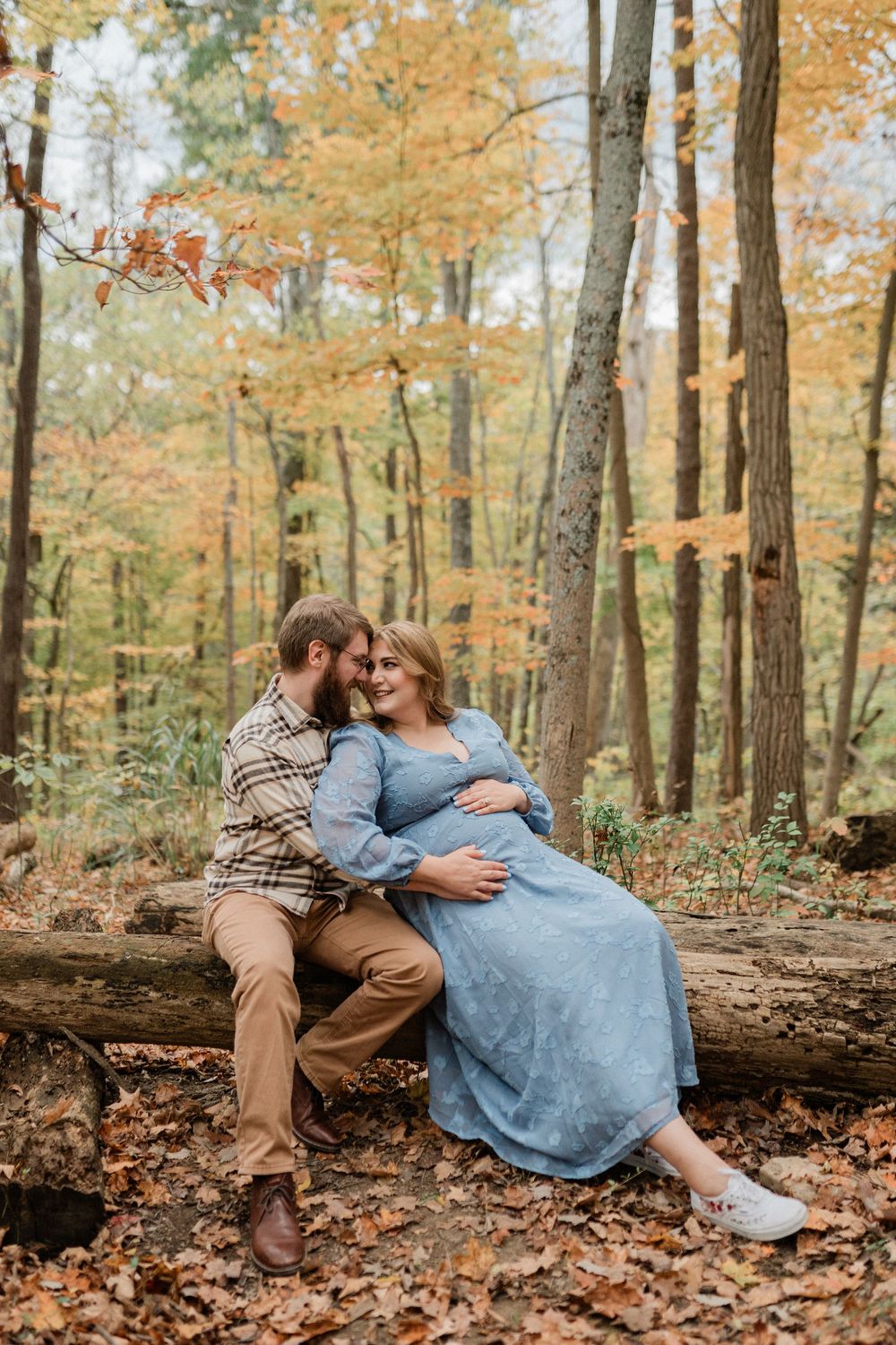 A couple sits together on a fallen log surrounded by autumn foliage in a forest setting.