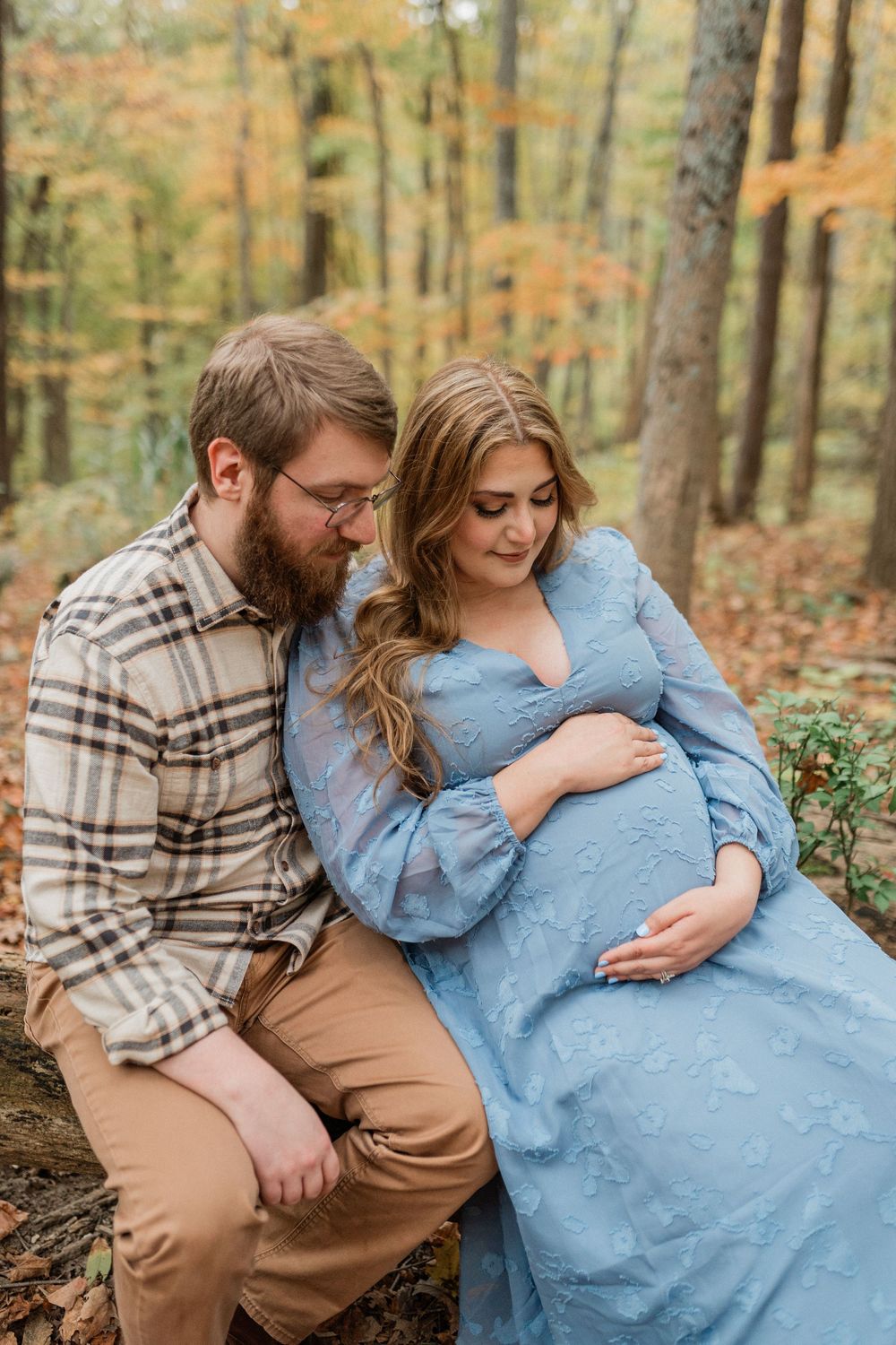 A couple sits together on a log in the autumn forest, sharing an intimate moment in casual attire.