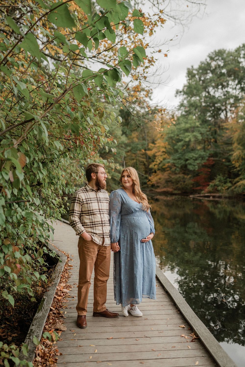 A couple stands together on a wooden dock surrounded by autumn foliage and reflective water.