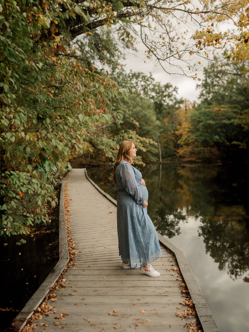 A wooden boardwalk extends over calm water with fall trees creating a peaceful autumn scene.