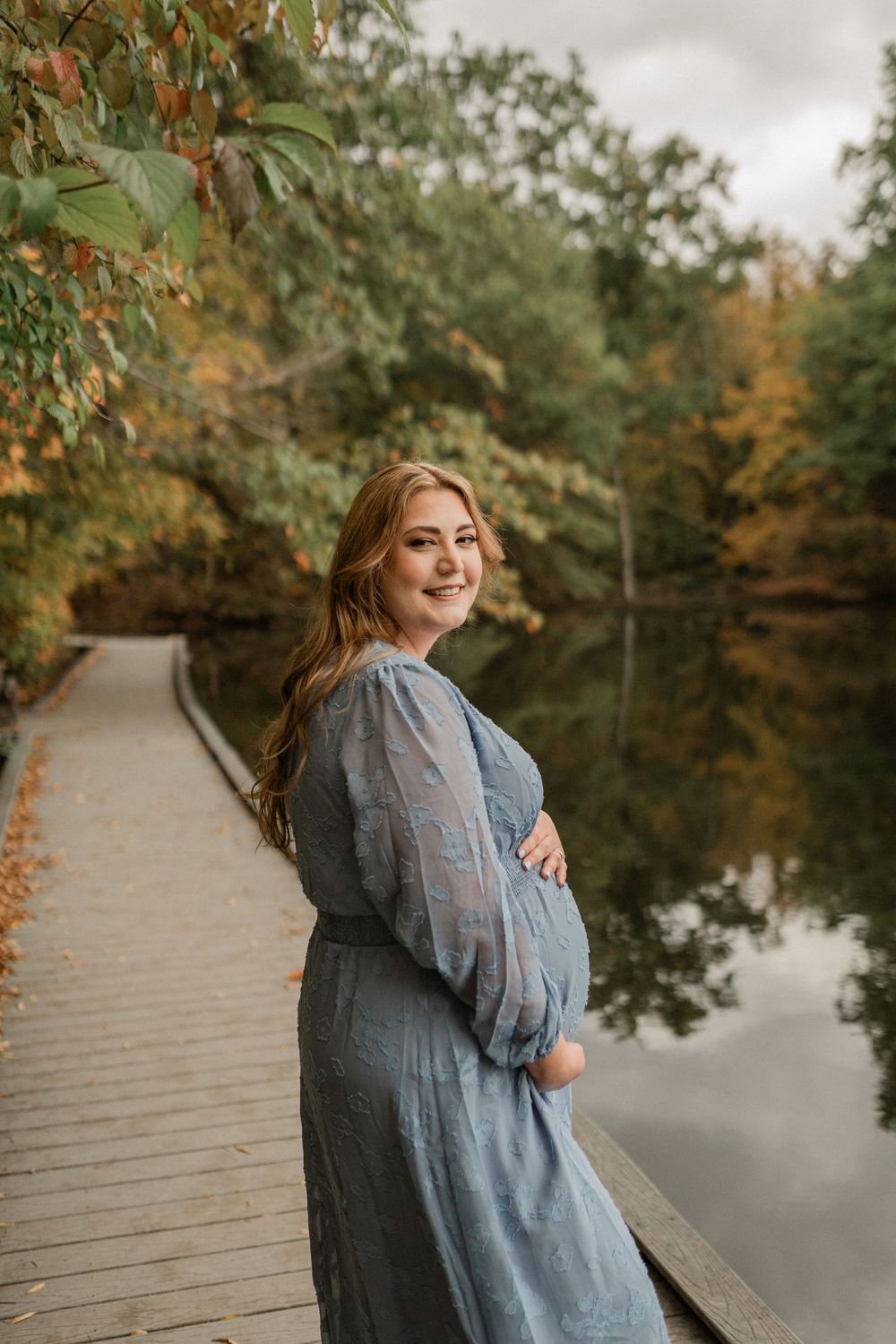 A woman in a blue dress poses on a wooden dock during an autumn maternity photoshoot.