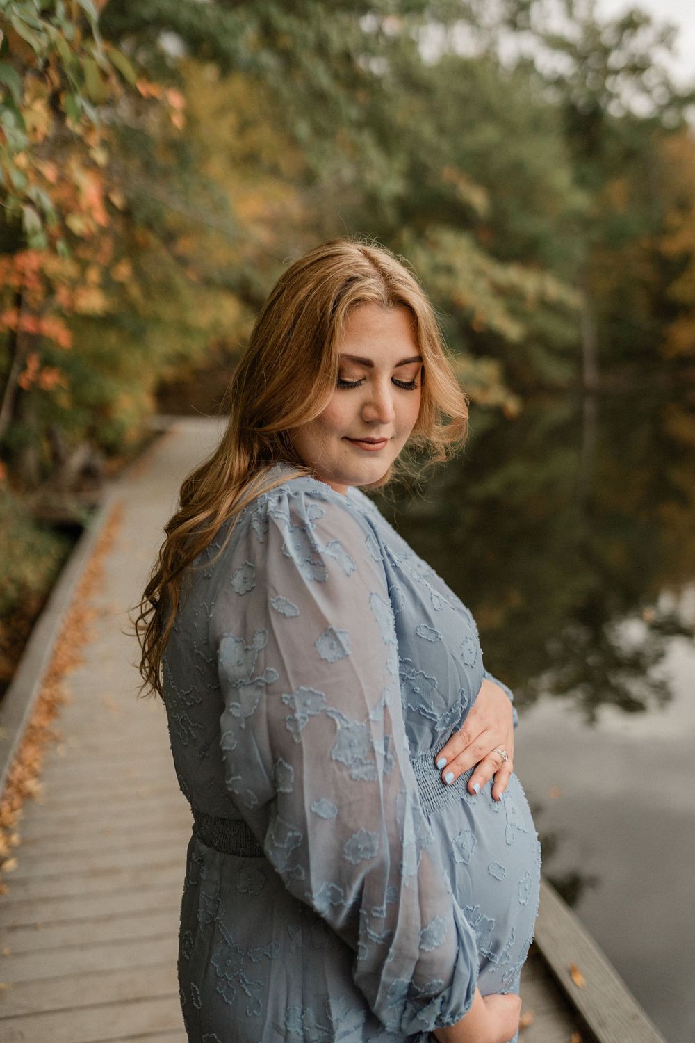 A woman in a blue lace dress stands on a wooden boardwalk during autumn, surrounded by fall foliage.
