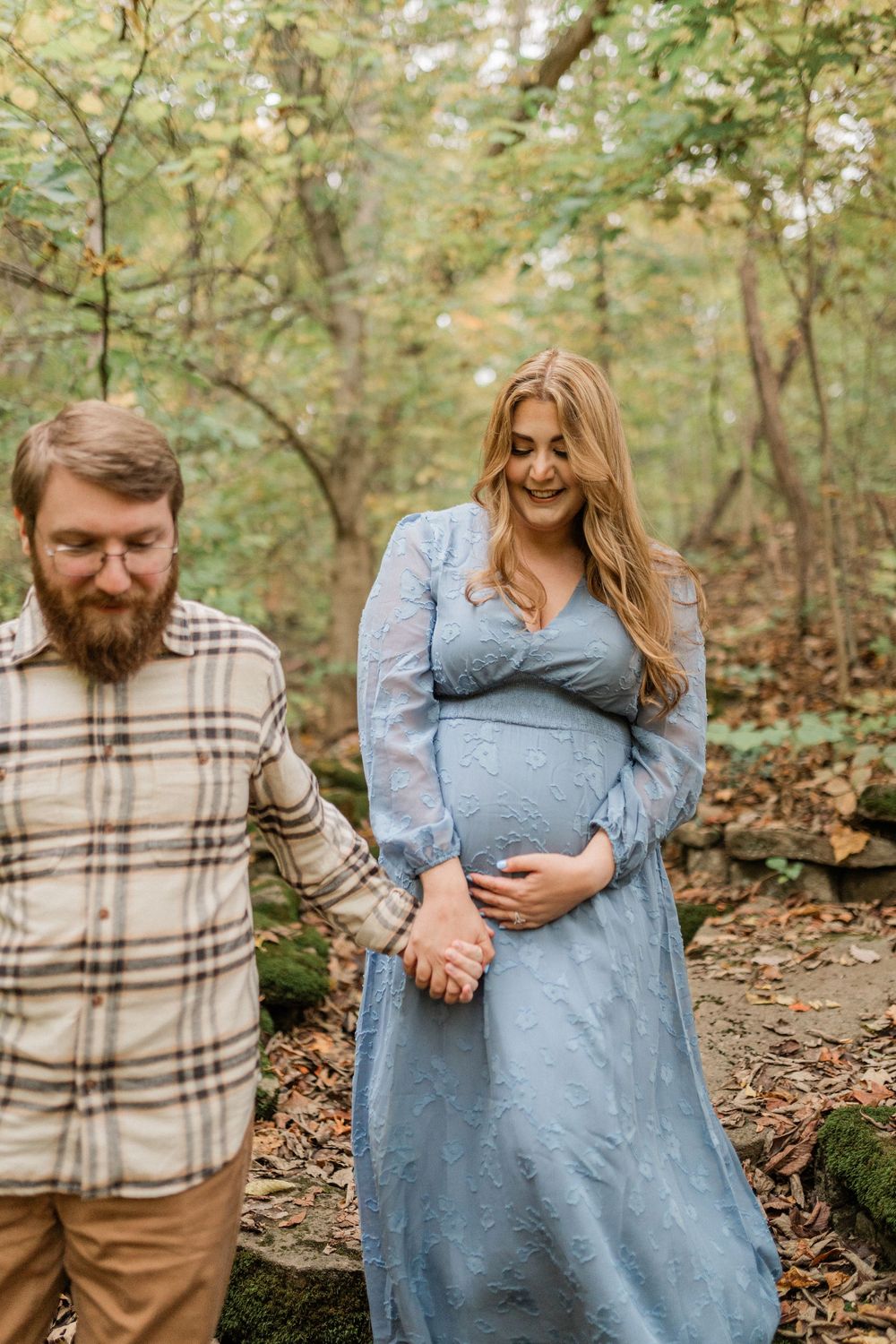 A couple in autumn woods share a tender moment as they walk together, wearing plaid and blue clothing.
