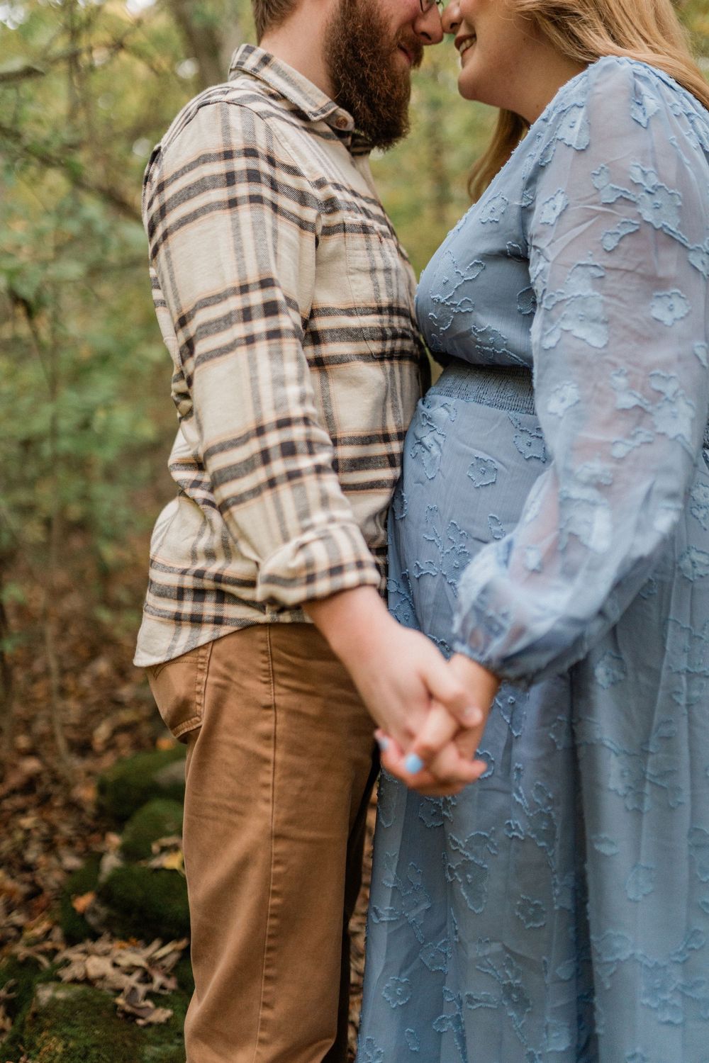A couple holds hands outdoors, wearing a plaid shirt and blue floral dress during a maternity photoshoot.
