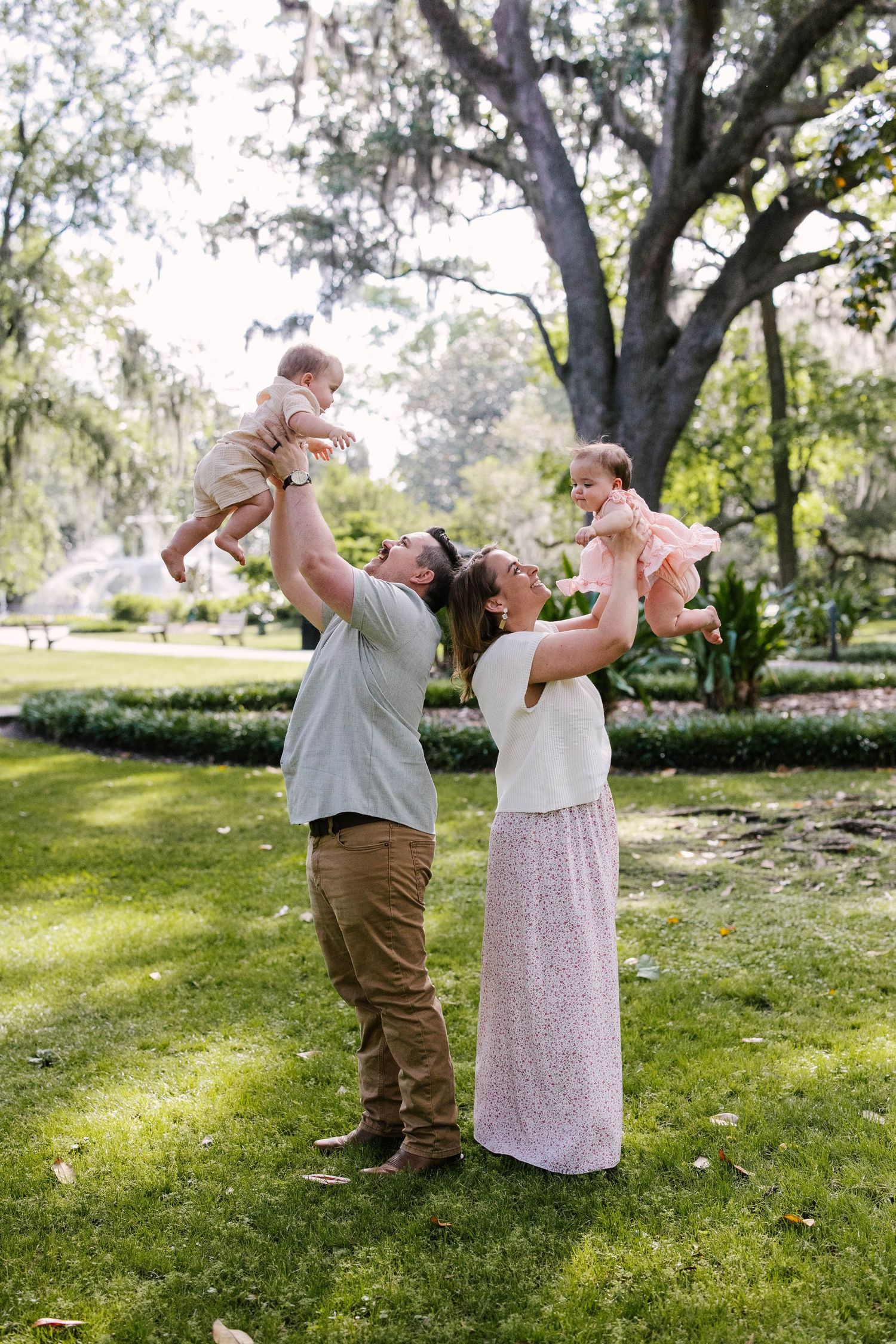 A family plays together on a sunny day, lifting children up high in a lush garden setting.