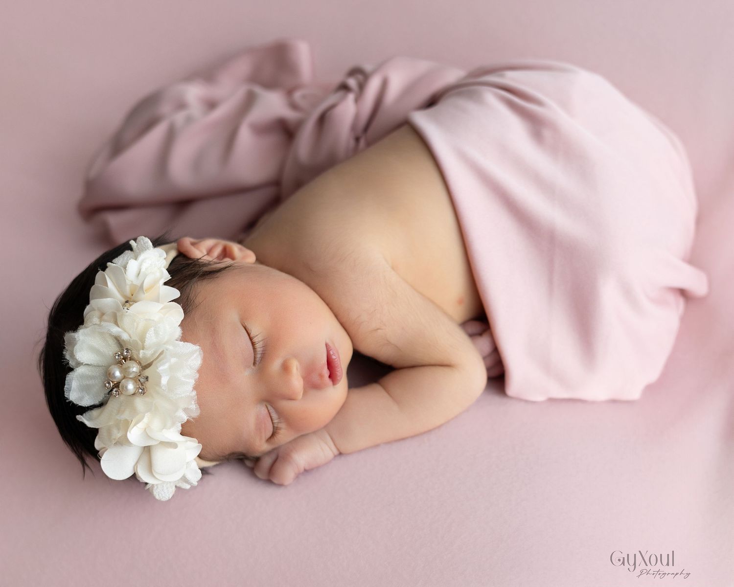A sleeping baby in pink swaddle blanket with white flower prop on pink background in newborn photography series.