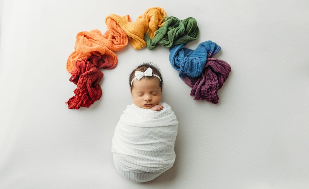 Newborn baby sleeping surrounded by rainbow colored fabric swatches.