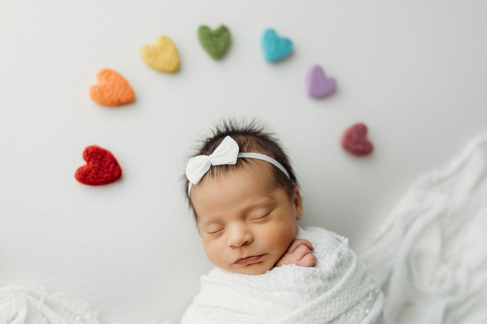 Sleeping newborn baby wrapped in white on light background with colorful heart decorations.