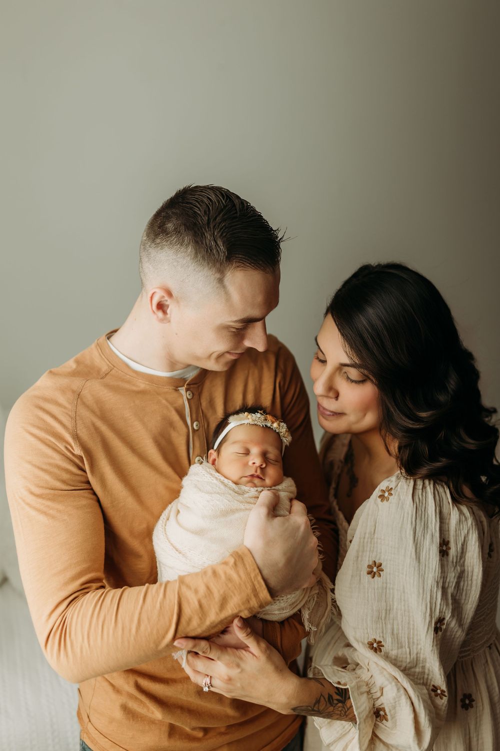 Family portrait with parents holding newborn baby against neutral wall.