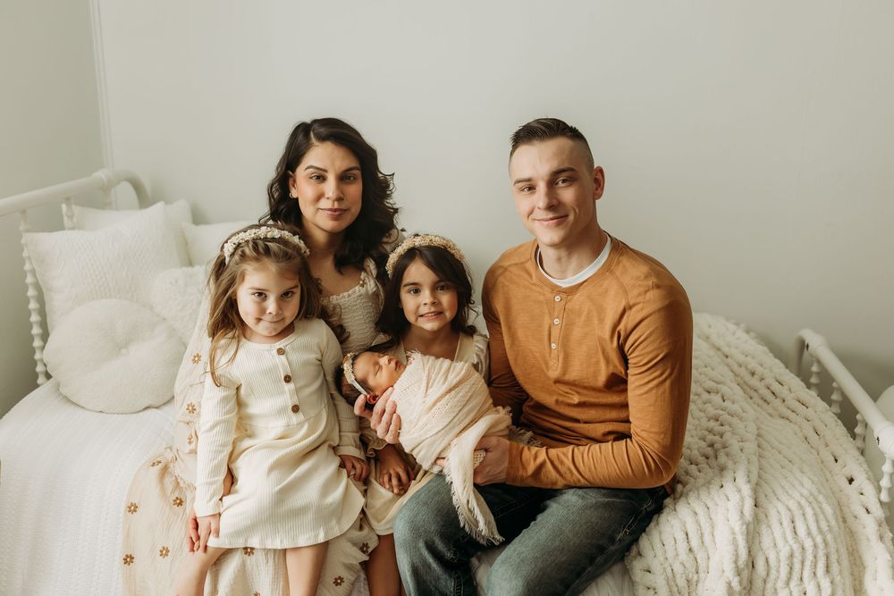 Family wearing coordinated neutral tones sits together on white bedding for an intimate lifestyle photoshoot.