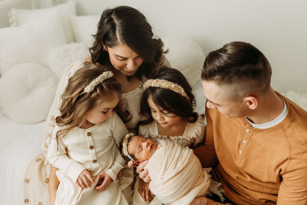 Family wearing neutral colored clothing gathers together on white couch to look at newborn baby.
