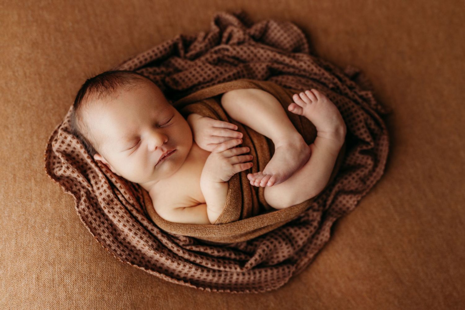 A series of five sepia-toned photos showing a sleeping newborn baby nestled in brown knit blankets.