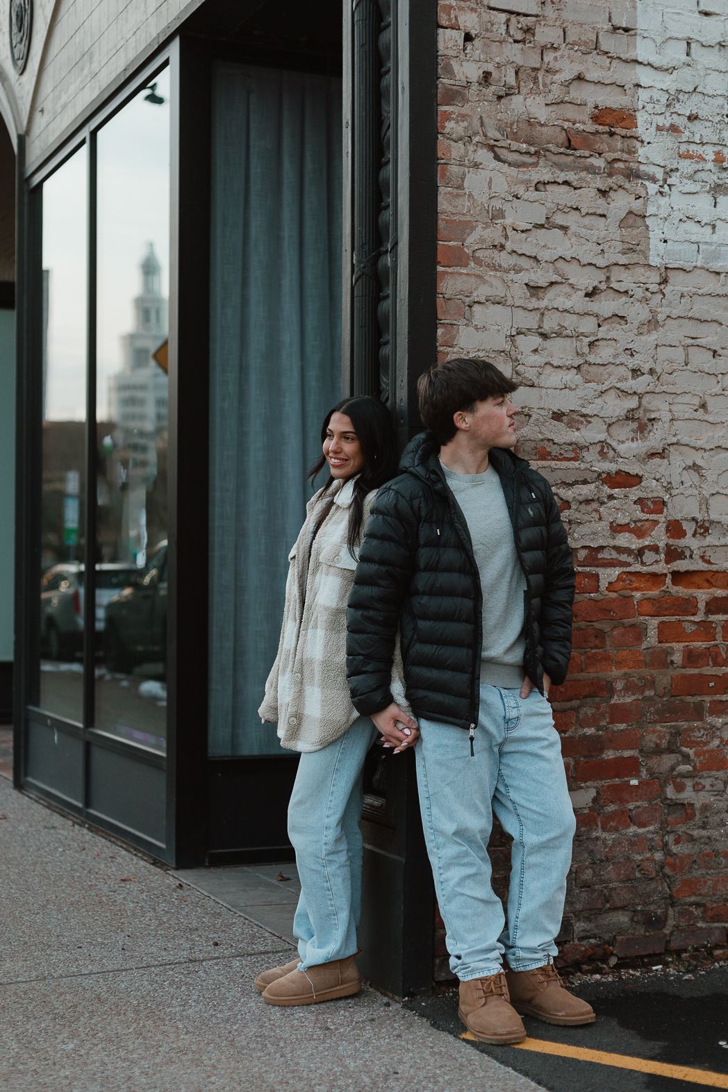 A couple in matching light denim and black puffer vests stand together against an exposed brick wall.