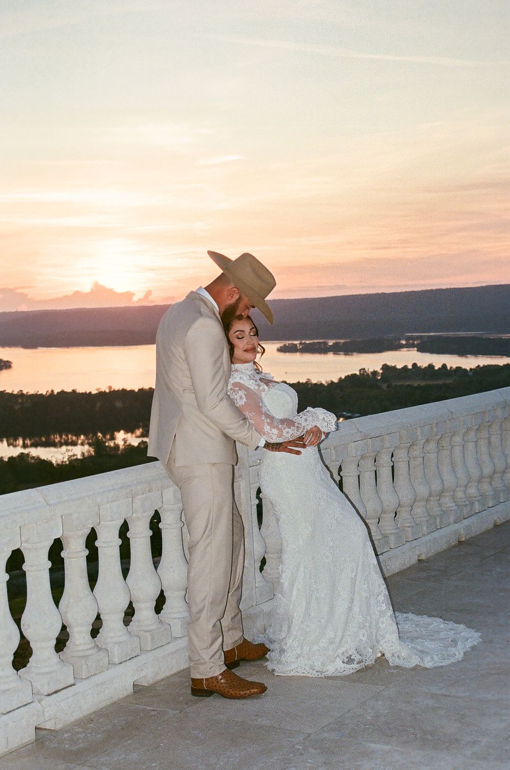 Bride and groom sharing intimate sunset moment on white balcony overlooking lake in western attire.