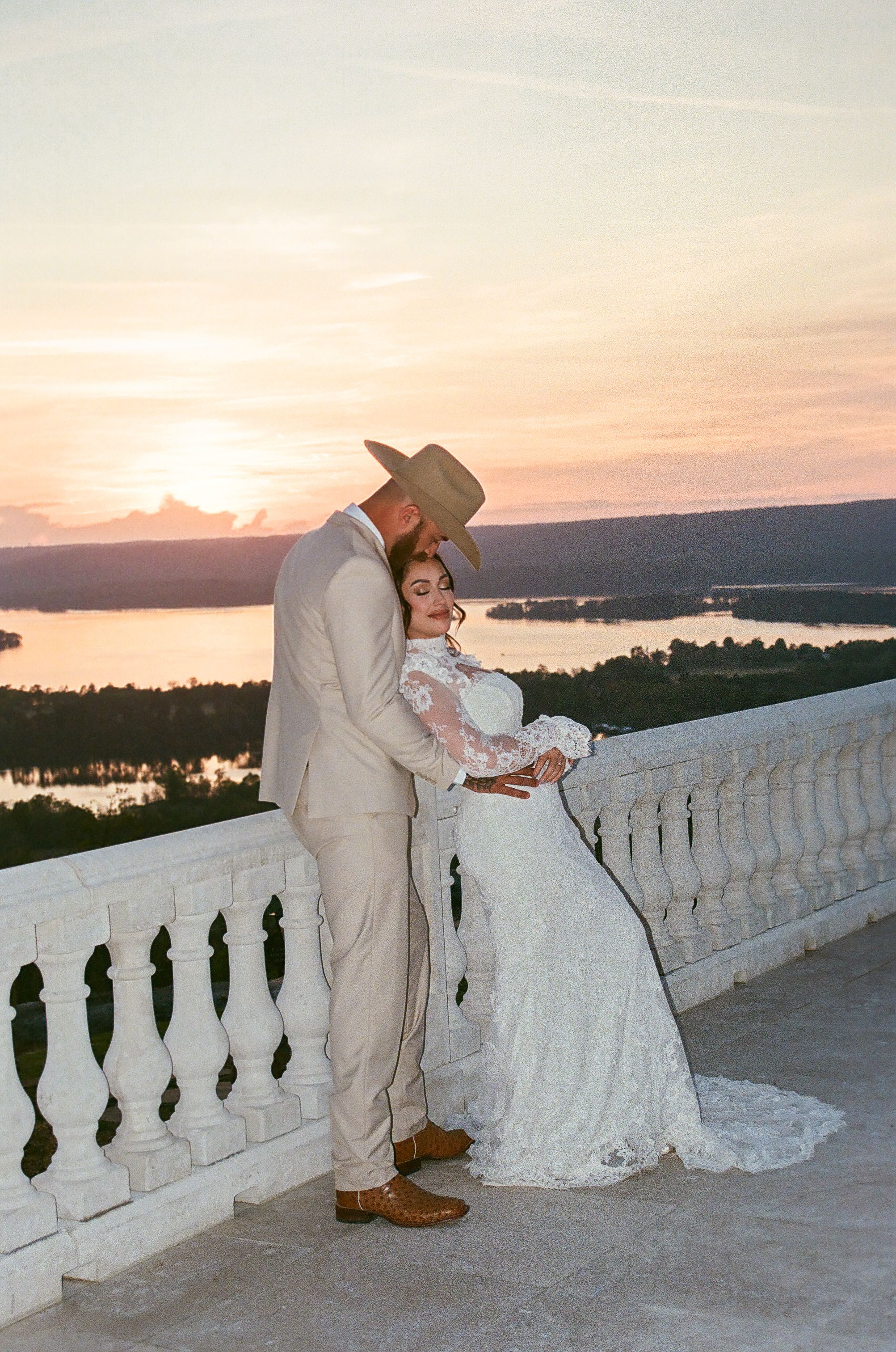 Bride and groom sharing intimate sunset moment on white balcony overlooking lake in western attire.