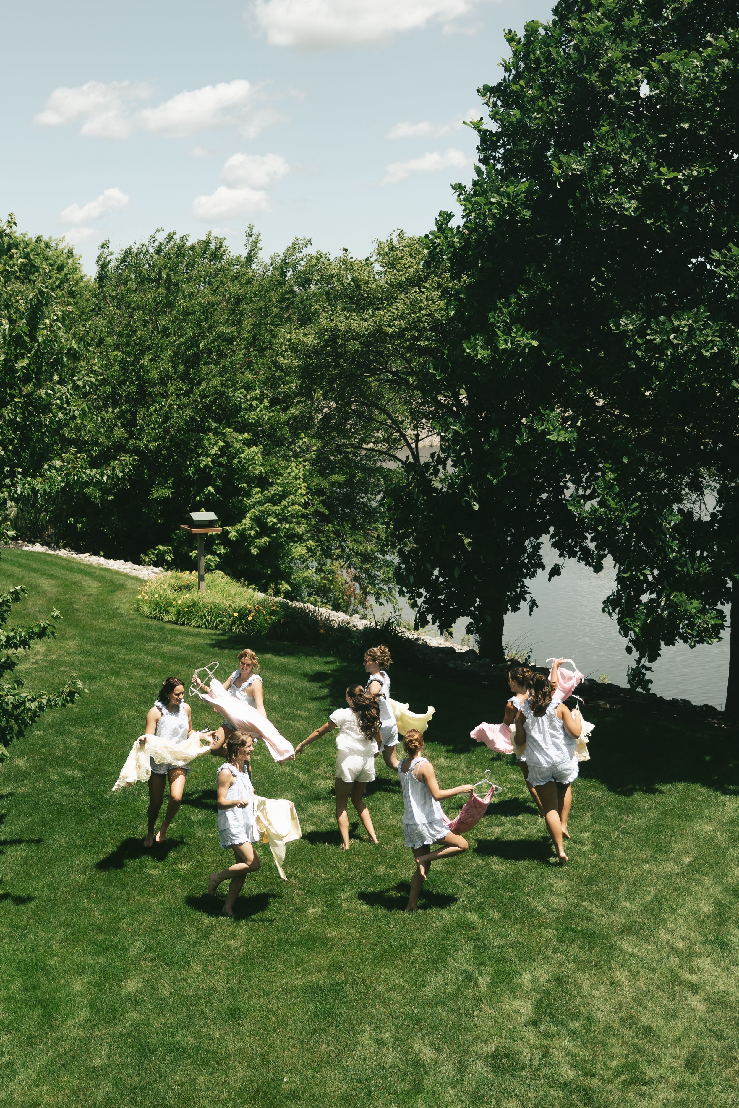 Group of women in white dresses joyfully running on lush green lawn surrounded by trees near a lake on a sunny day.