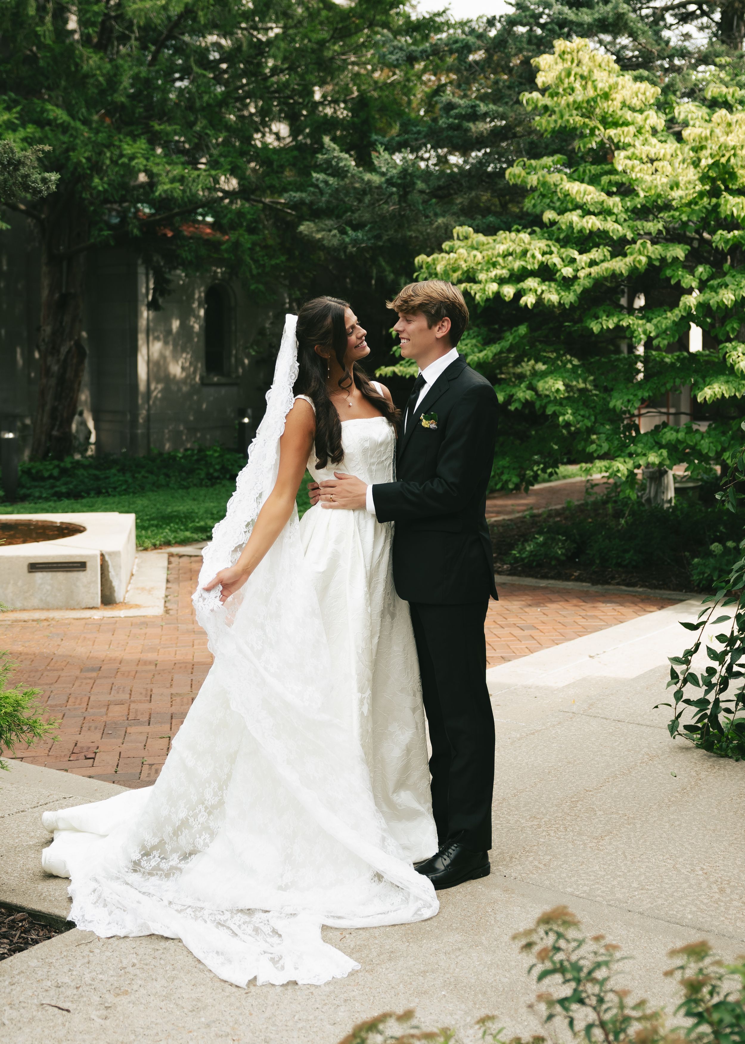 Bride in white gown with long veil and groom in black tuxedo posing together outdoors on brick path surrounded by trees.