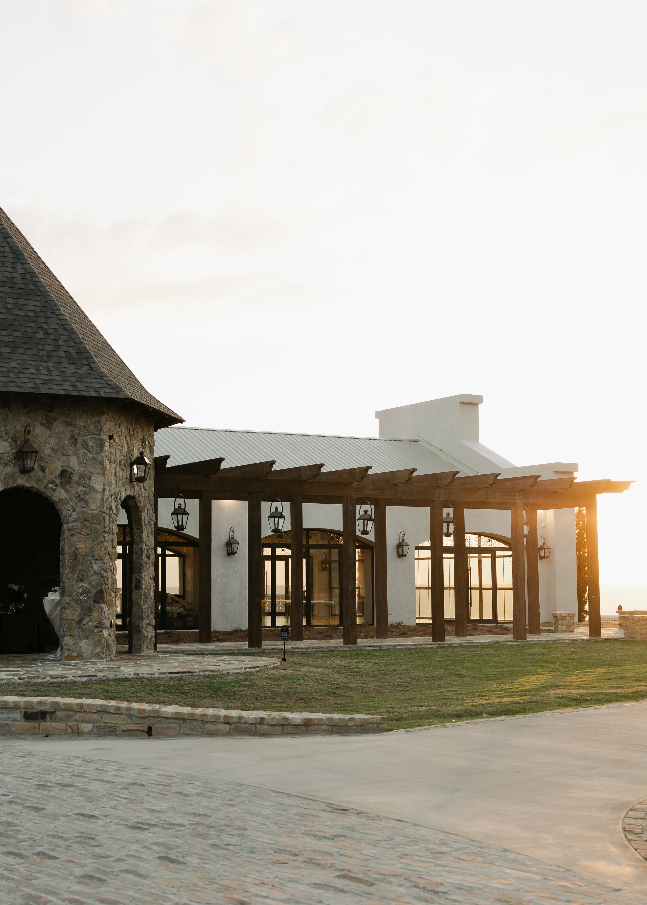 Elegant stone and white stucco event venue with wooden pergola and lanterns at golden hour sunset.