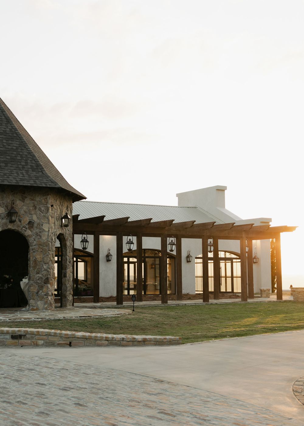 Elegant stone and white stucco event venue with wooden pergola and lanterns at golden hour sunset.