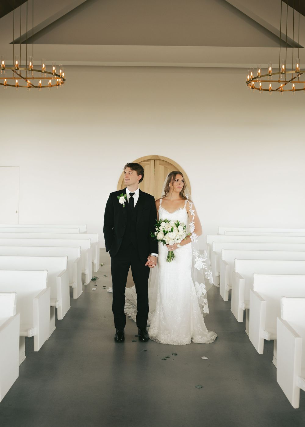Bride and groom hold hands in a white chapel aisle, she in lace gown with bouquet, he in black tuxedo.