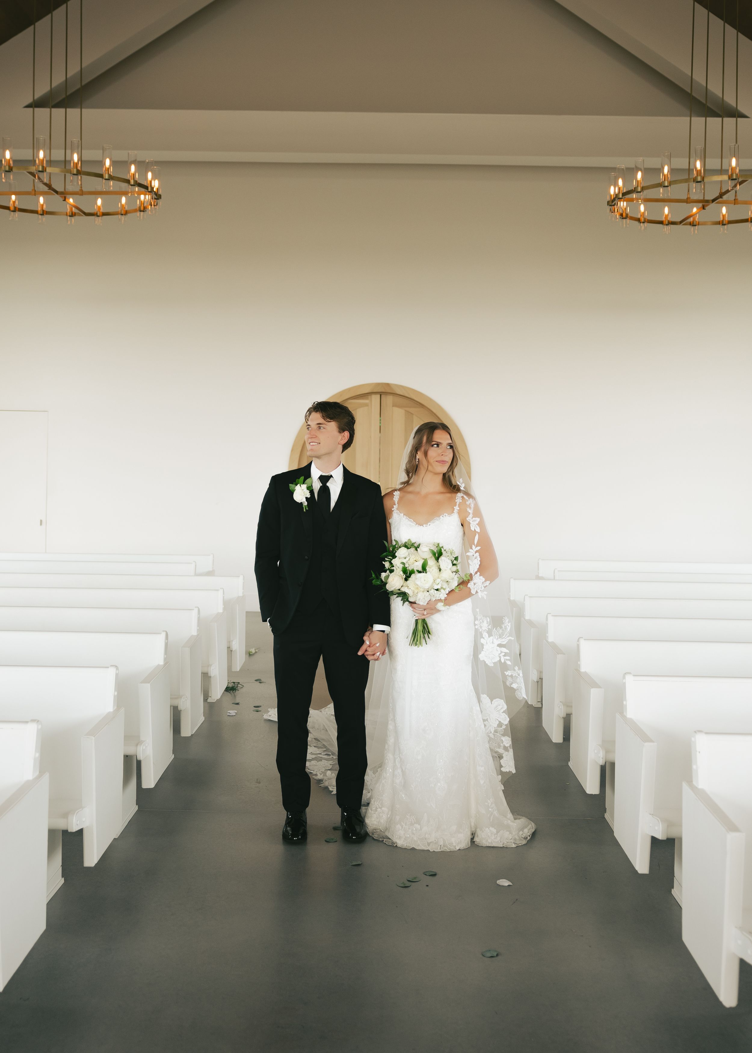 Bride and groom hold hands in a white chapel aisle, she in lace gown with bouquet, he in black tuxedo.