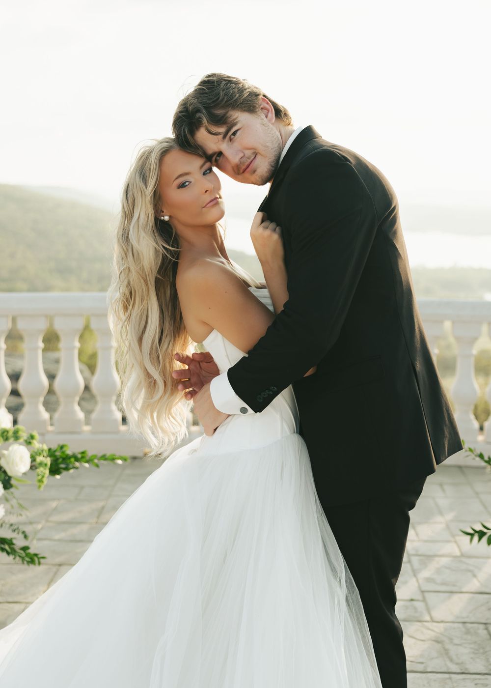 Romantic bride and groom embracing on outdoor terrace with scenic lake view at golden hour.