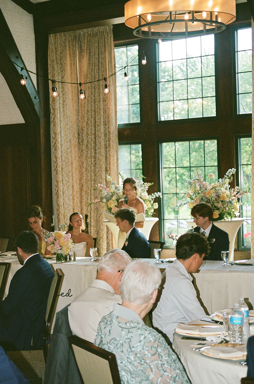 Elegant wedding reception in a rustic venue with floral arrangements, string lights, and guests seated at white-draped tables.