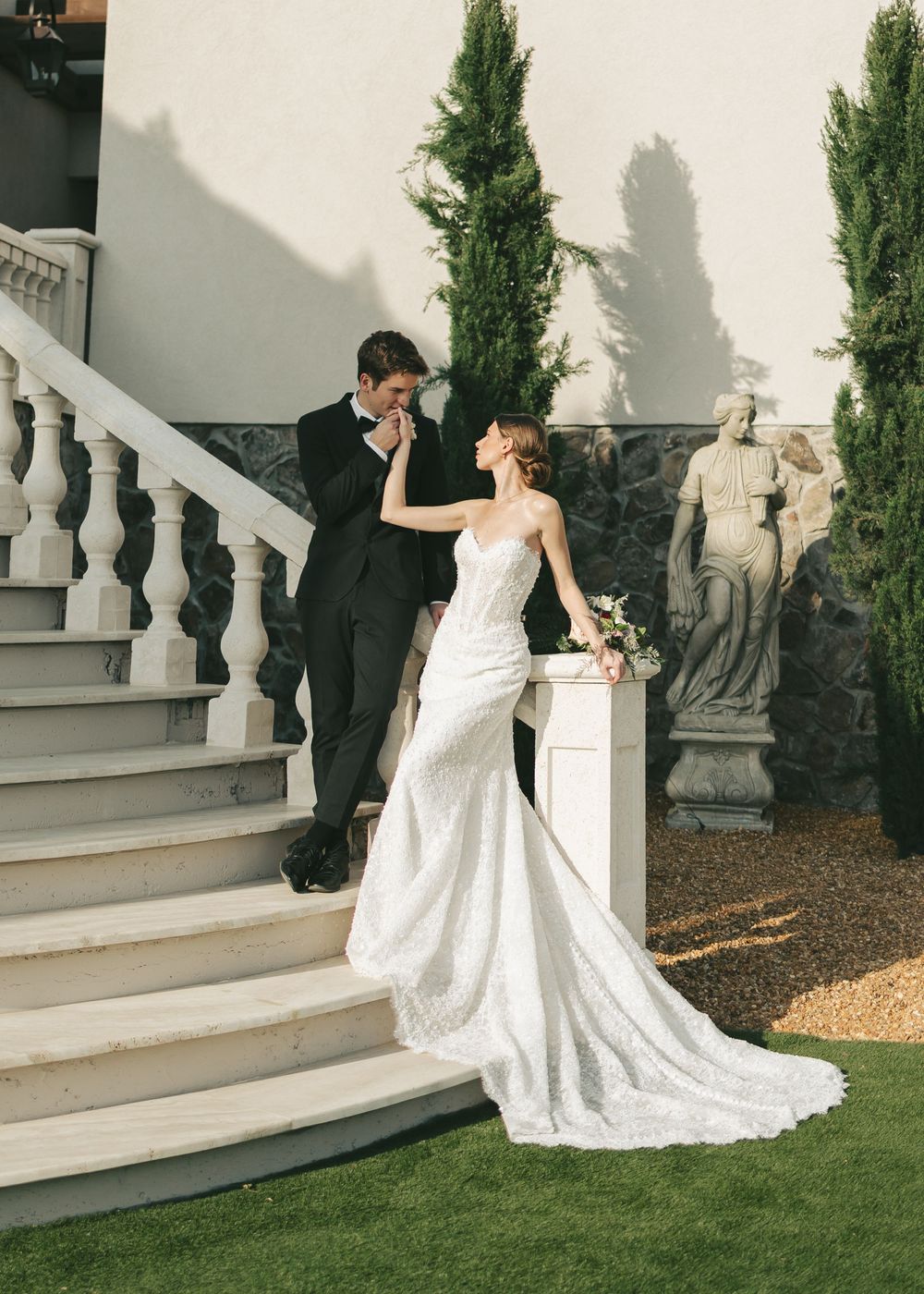 A groom in a black tuxedo kisses his bride's hand on elegant white stone stairs, as she wears a fitted lace wedding gown.