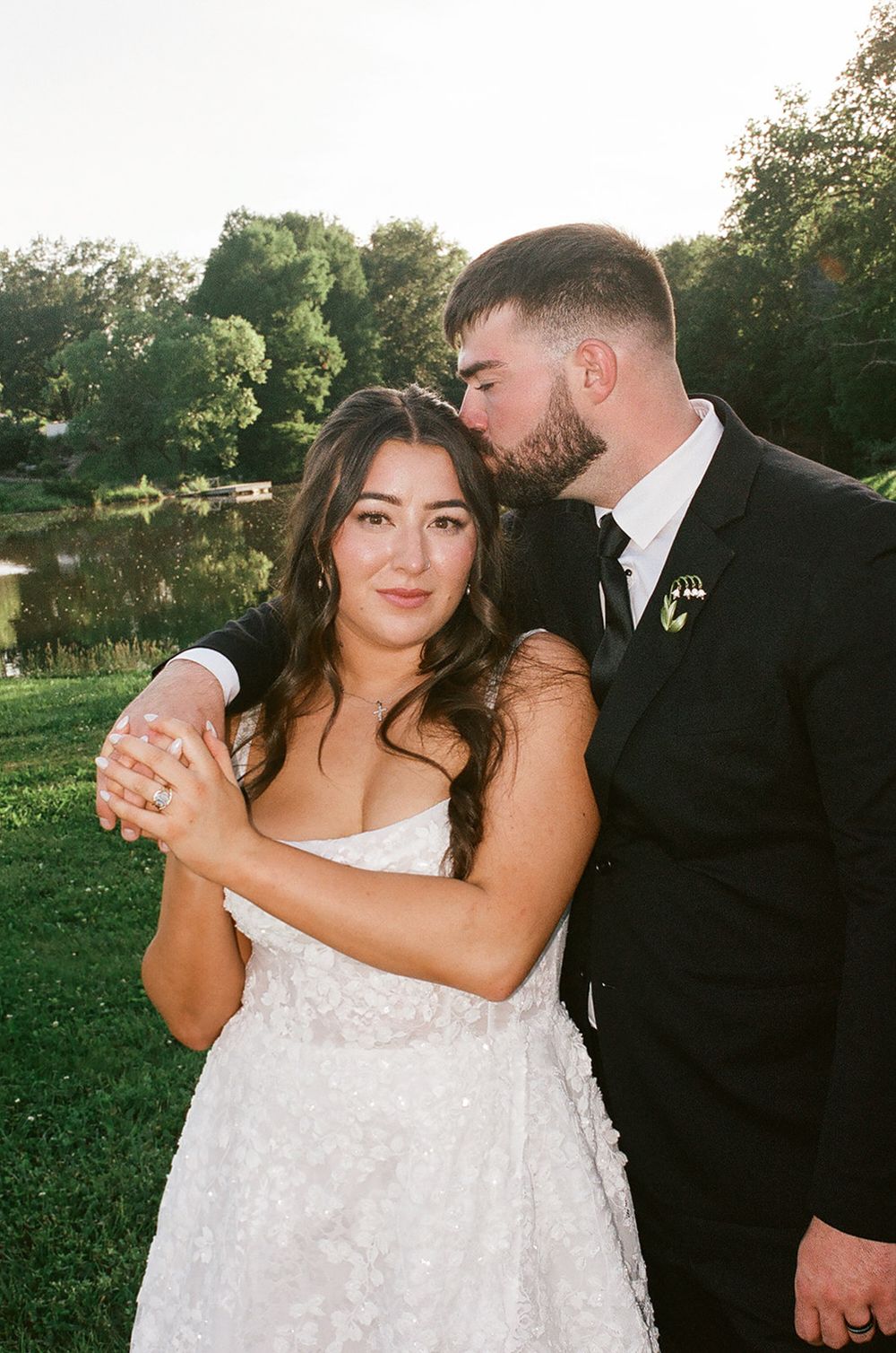 Groom in black suit kisses bride on cheek while embracing outdoors near pond at golden hour.
