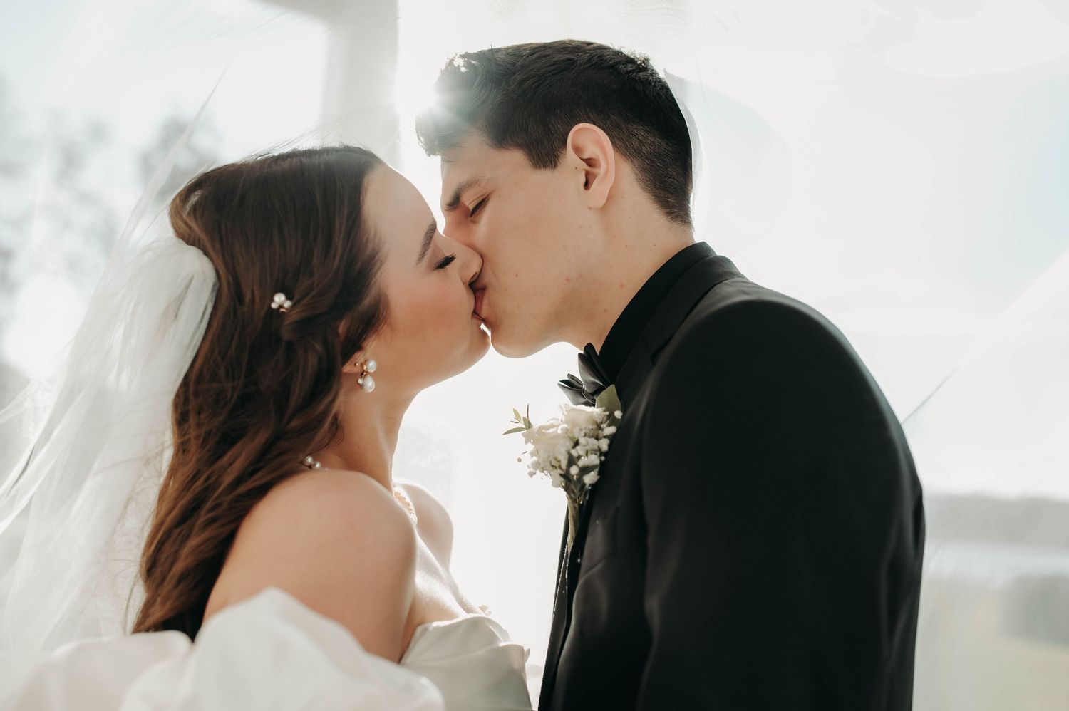 Bride and groom sharing first kiss in sunlit venue, bride in off-shoulder gown with veil and pearl pins.