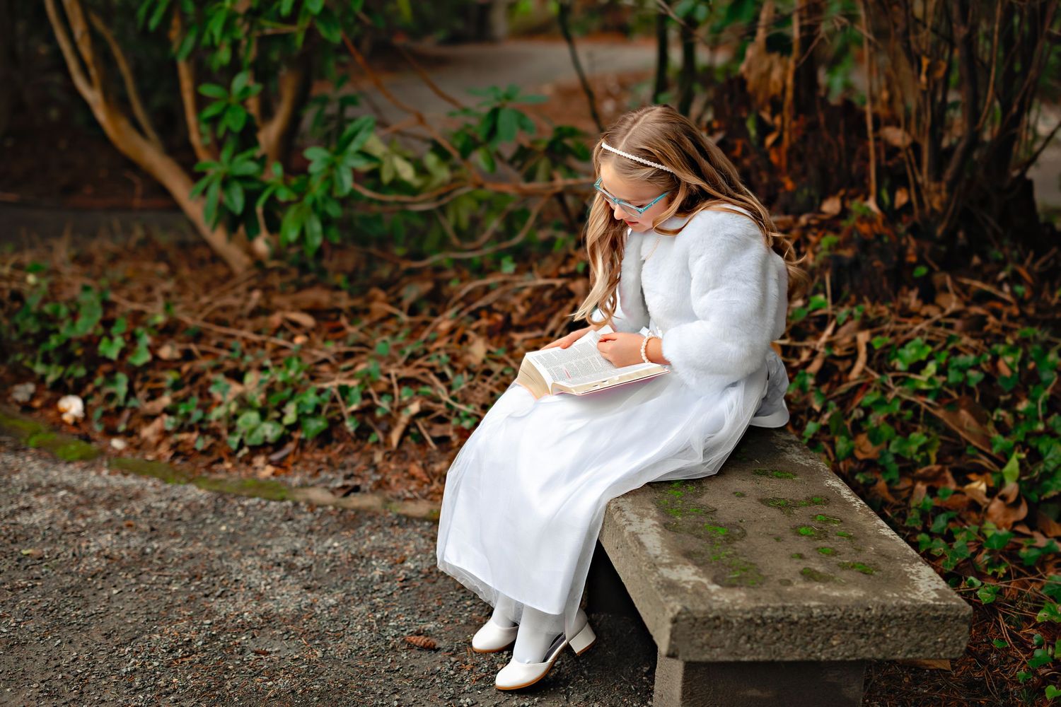 A figure in a flowing white dress and brown shoes sits on a stone bench in a garden setting with autumn leaves.