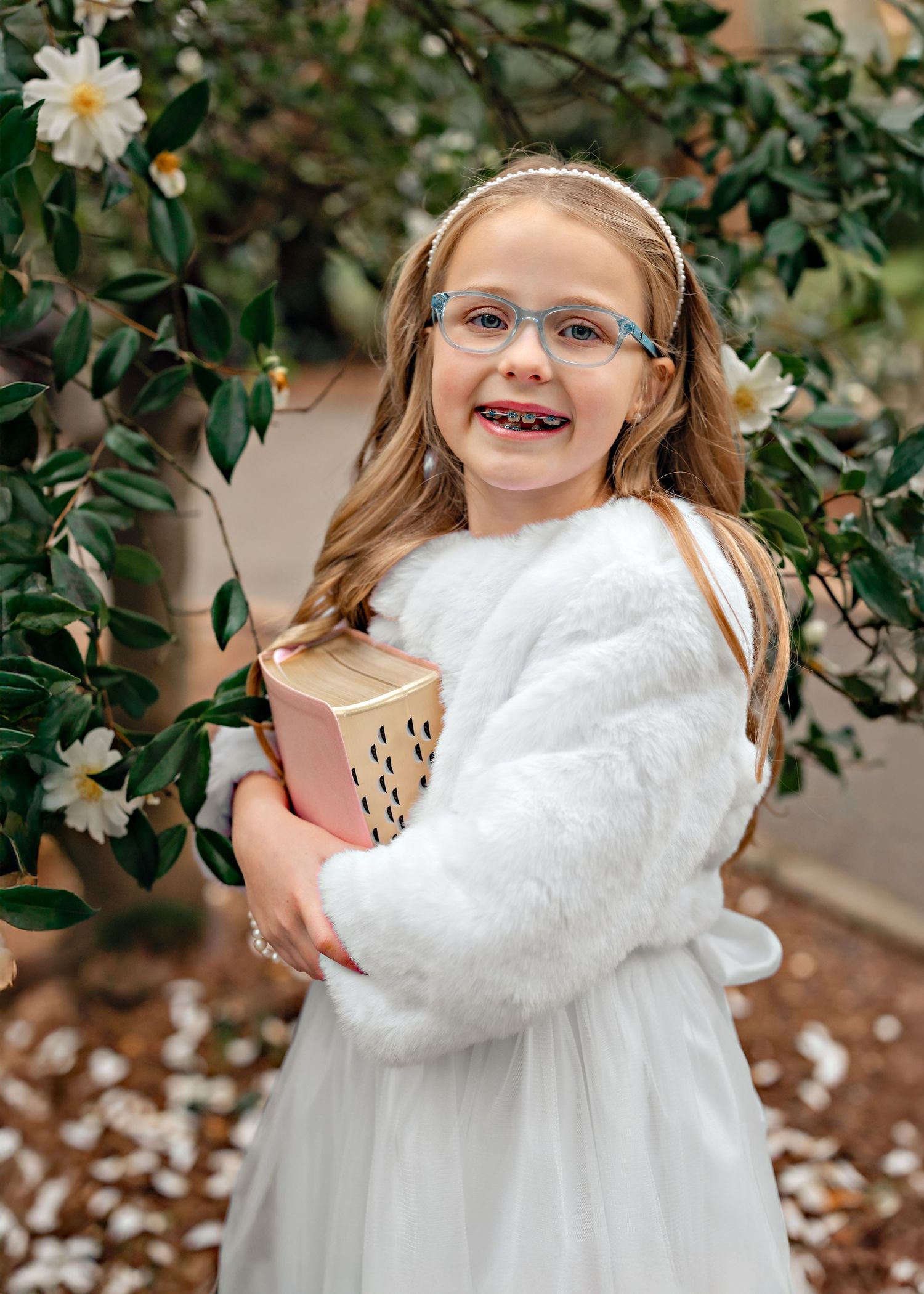 A child in a white sweater and glasses hugs a plush toy while standing among daisy flowers and greenery.